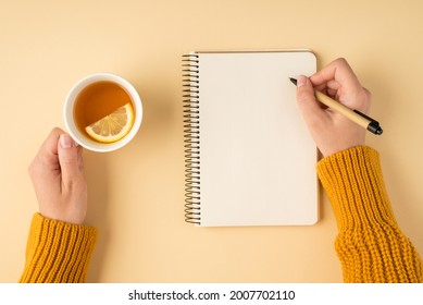 First person top view photo of female hands in yellow pullover writing in spiral planner and holding white cup of tea with lemon slice on isolated pastel orange background with copyspace