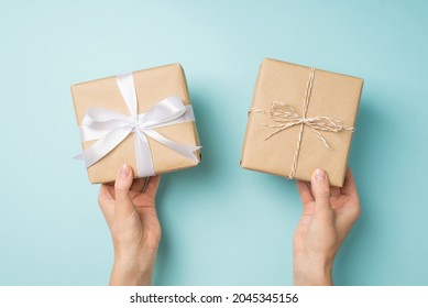 First Person Top View Photo Of Female Hands Holding Two Craft Paper Gift Boxes With Twine Bow And White Satin Ribbon Bow On Isolated Pastel Blue Background