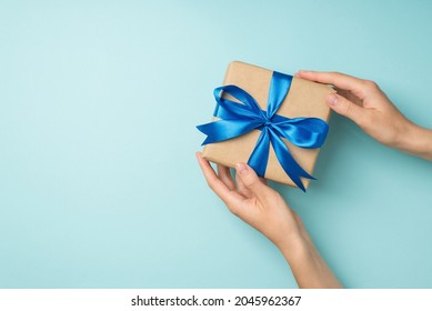 First person top view photo of woman's hands holding craft paper giftbox with blue satin ribbon bow on isolated pastel blue background with copyspace - Powered by Shutterstock