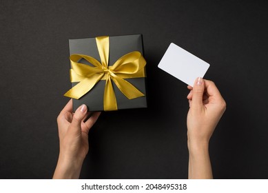 First person top view photo of hands holding black giftbox with golden satin ribbon bow and white plastic card on isolated black background with blank space