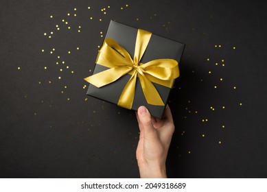 First person top view photo of hand holding black giftbox with yellow satin ribbon bow over shiny golden sequins on isolated black background with empty space - Shutterstock ID 2049318689