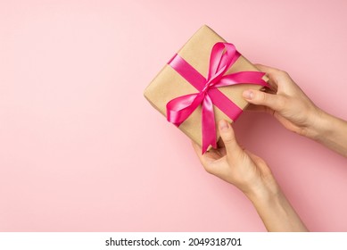 First person top view photo of female hands giving stylish craft paper giftbox with pink satin ribbon bow on isolated pastel pink background with empty space