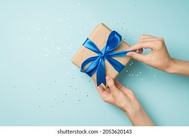 First person top view photo of hands unpacking craft paper gift box with blue satin ribbon bow over shiny sequins on isolated pastel blue background with blank space - Shutterstock ID 2053620143