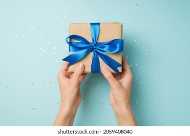 First person top view photo of hands holding craft paper giftbox with vivid blue ribbon bow over shiny sequins on isolated pastel blue background - Shutterstock ID 2059408040