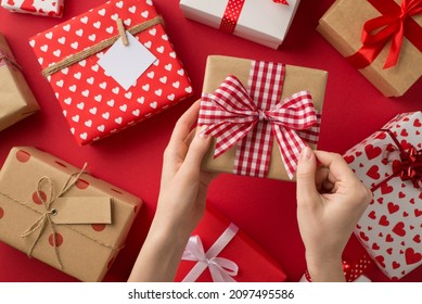 First person top view photo of saint valentine's day decorations young woman's hands untying checkered ribbon bow on kraft paper giftbox over present boxes on isolated red background