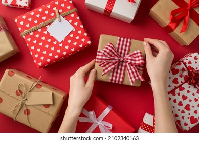 First person top view photo of st valentine's day decorations presents and female hands taking kraft paper giftbox with checkered ribbon bow on isolated red background