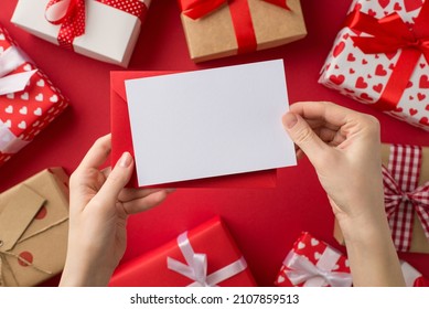 First person top view photo of saint valentine's day decor young female hands holding red envelope and card over present boxes on isolated red background with blank space