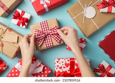 First person top view photo of st valentine's day decor presents and woman's hands tying checkered ribbon bow on craft paper giftbox on isolated pastel blue background