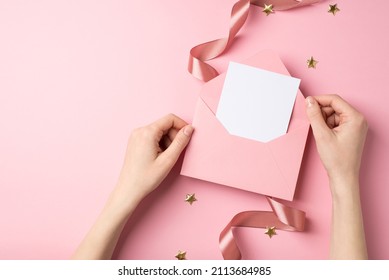First person top view photo of valentine's day decor woman's hands holding open pink envelope with paper card golden stars and pink silk curly ribbon on isolated pastel pink background with copyspace