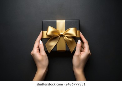 First person top view photo of hand holding black giftbox with yellow satin ribbon bow over shiny golden sequins on isolated black background with empty space
