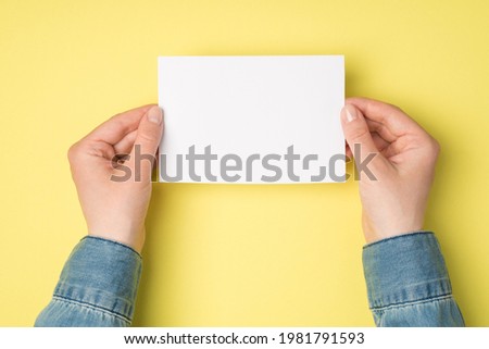 First person top view photo of female hands holding white paper card on isolated yellow background with blank space
