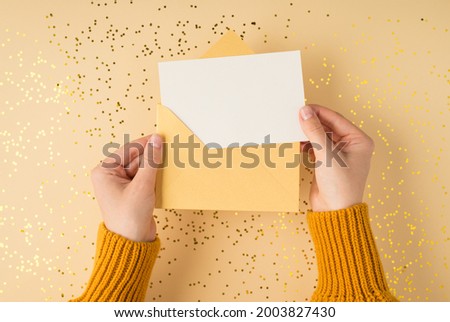 First person top view photo of female hands in yellow pullover holding open pastel yellow envelope with white card over scattered golden sequins on isolated pastel orange background with blank space