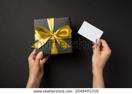 First person top view photo of hands holding black giftbox with golden satin ribbon bow and white plastic card on isolated black background with blank space