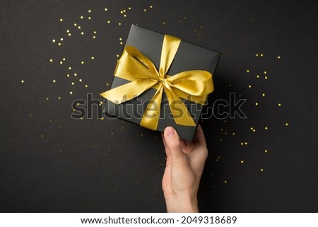 First person top view photo of hand holding black giftbox with yellow satin ribbon bow over shiny golden sequins on isolated black background with empty space