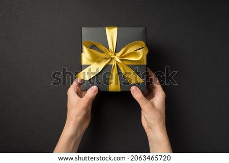 First person top view photo of hands giving black giftbox with golden satin ribbon bow on isolated black background