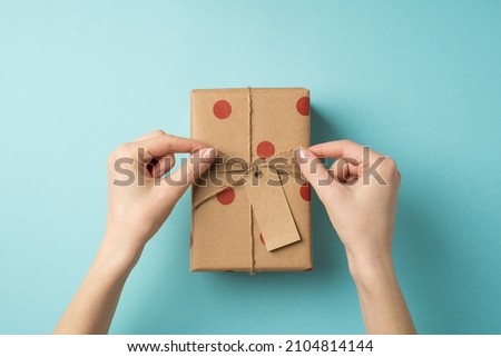 First person top view photo of valentine's day decorations female hands tying twine bow on craft paper gift box with polka dot pattern and pricetag on isolated pastel blue background with blank space
