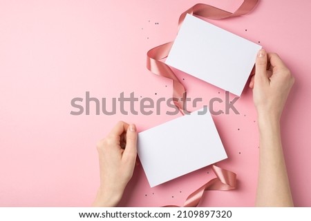 First person top view photo of valentine's day decorations girl's hands holding two paper sheets sequins and silk curly ribbon on isolated pastel pink background with copyspace