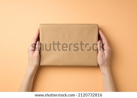 First person top view photo of young woman's hands holding kraft paper giftbox on isolated beige background with empty space