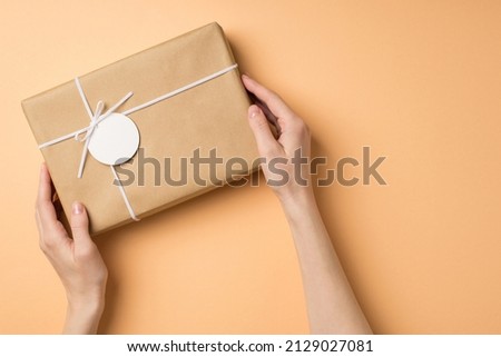 First person top view photo of st valentine's day decorations female hands holding kraft paper giftbox with bow and tag on isolated beige background with copyspace