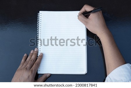First person top view photo of male hands in black pullover writing in spiral notepad. Black table background with copyspace