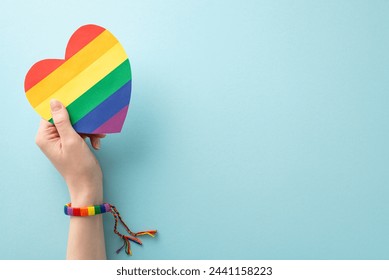 First person top-down view shot of a woman's hand in symbolic bracelet holding a rainbow heart-shaped card on a pastel blue background with an empty space for text or advertising - Powered by Shutterstock