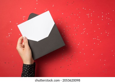 First person view above photo of woman hand holding black envelope with card inside isolated on the red background with confetti
