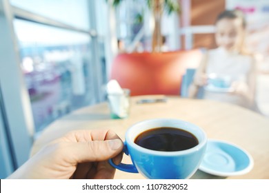 First Person View On Blue Cup Of Coffee With Woman On Background. Couple Sitting In Coffee Shop And Having Coffee