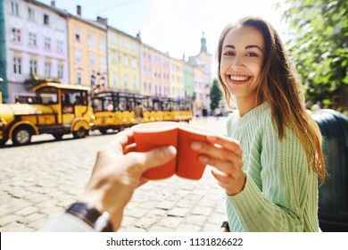 First Person View On A Woman In Green Sweater Drinking Coffee From Small Paper Cups With Boyfriend. Attractive Happy Smiling Woman Sitting In The Center Of Old City With Small Yellow Tourist Train On