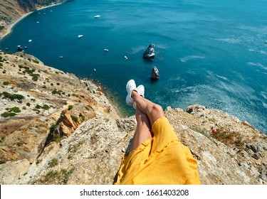 first person view of tanned woman legs in yellow dress sitting on on cliff edge with amazing nature view of seascape with high limestone cliff over blue sea.
