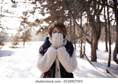 first snow. a woman stands against the backdrop of a snowy forest, covering her face with knitted mittens. She's dressed warmly in a shearling jacket and plaid scarf, creating a cozy winter vibe.