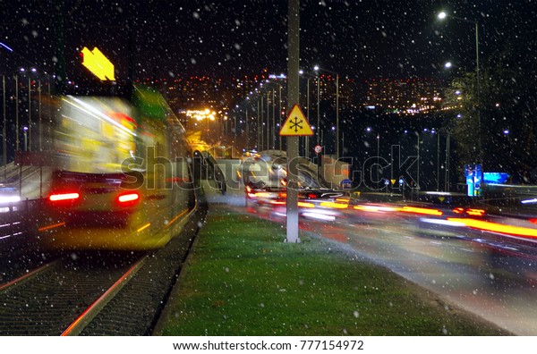 First snowfall in\
city by night. Blurred lights cars and tramway on town street with\
snowflakes in first plan.