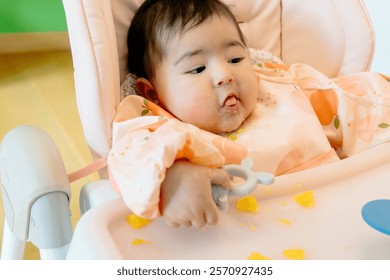 First solid feeding of cute 6 month old baby sitting in high chair dirty and messy, spilled food. Feeding problems after weaning.