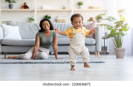 First Steps. Adorable Black Infant Child Walking In Living Room At Home, Cute African American Toddler Boy Stepping On Floor And Looking At Camera, His Proud Mother Smiling On Background, Free Space