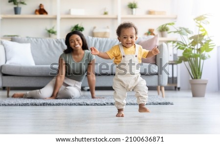 First Steps. Adorable Black Infant Child Walking In Living Room At Home, Cute African American Toddler Boy Stepping On Floor And Looking At Camera, His Proud Mother Smiling On Background, Free Space