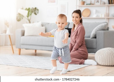 First Steps. Adorable Smiling Baby Boy Learning How To Walk At Home, Happy Mom Proud Of Her Little Child Sitting On Background, Copy Space