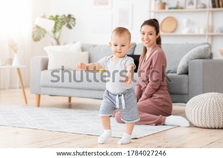 First Steps. Adorable smiling baby boy learning how to walk at home, happy mom proud of her little child sitting on background, copy space