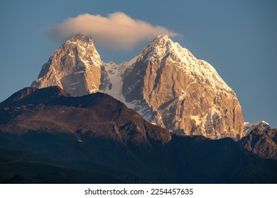 First sunrise reaching the peaks of Ushba in Caucasus, Georgia. Cloudless sky above the high and snow-capped mountains. The hills below are shrouded with shadows. Daybreak. High and desolate mountains - Shutterstock ID 2254457635