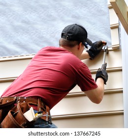 First time home buyer works to install siding on his new home.  He is hammering into place a sheet of siding.  He has on a red shirt and is holding hammer and nail.