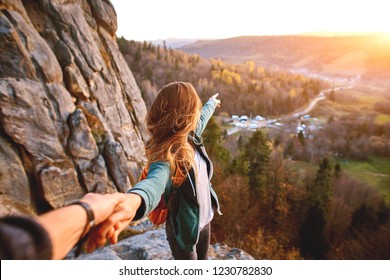 first view of woman hiker with backpack stands on edge of cliff against background of sunrise and extends a hand
