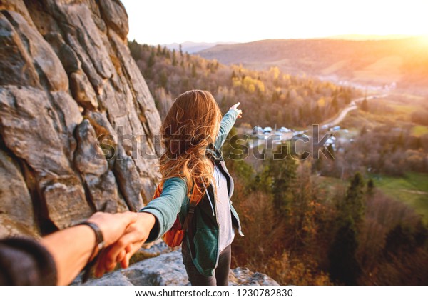 first view of\
woman hiker with backpack stands on edge of cliff against\
background of sunrise and extends a\
hand