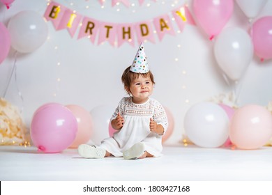 First year's birthday. happy little girl in white dress and festive hat sits in background with garlands and pink balloons, celebrating her first birthday. Decoration birthday. copy space.