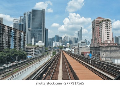 A first-person perspective looking down the elevated LRT railway tracks heading toward the Kuala Lumpur skyline, Malaysia. The symmetrical tracks create a strong leading line toward a dense cluster of modern skyscrapers and glass office towers. The scene captures the contrast between urban transit infrastructure and the city's architectural growth under a bright, cloudy sky, showcasing the pulse of a major Southeast Asian metropolis.