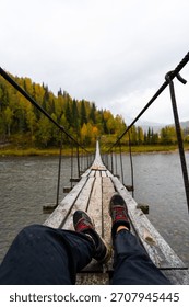 First-person point of view looking down the length of a wooden suspension bridge over a river, with the traveler's legs and hiking boots in the foreground. Person present is Caucasian, adult