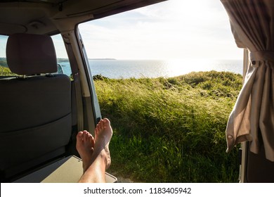 First-person View Of A Barefoot Man Relaxing Inside A Camper Van And Enjoying The View Over The Sea At Sunset Through The Open Sliding Door With Wild Grasses In The Foreground.