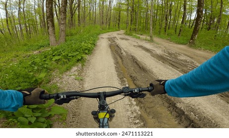 First-person view of a cyclist riding along a muddy forest path. The cyclist's gloved hands are gripping the handlebars of the bike. The uneven terrain and dense undergrowth add to the thrill - Powered by Shutterstock