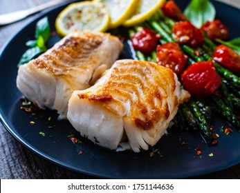 Fischgericht - Frittiertes Kabeljaufilet mit Spargel und Kirschtomaten auf schwarzem Teller auf Holztisch serviert 