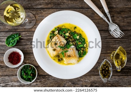 Fish dish - fried cod with spinach and capers in saffron sauce on wooden table 