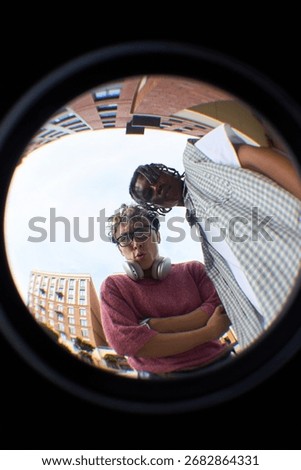 Fish eye view of two young men, Caucasian with curly hair and glasses, Black with braided hair, standing outdoors in urban setting, looking down into camera with serious expressions
