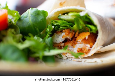 A Fish Finger Wrap With Salad On The Side. Food Is On A White Plate, In A Cafe. Salad Includes Cucumber, Lettuce, Rocket Leaves And Tomatoes.