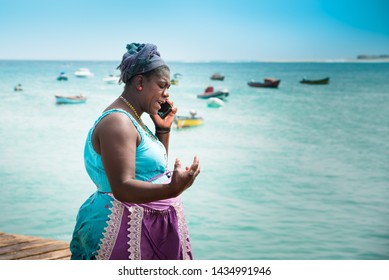 Fish Market, Santa Maria Pier, Sal, Cabo (Cape) Verde Islands - June 16, 2019: Older African Woman (fishmonger) In Colorful Working Clothes Talks Over Smartphone On Pier. Boats, Turquoise Sea Backdrop